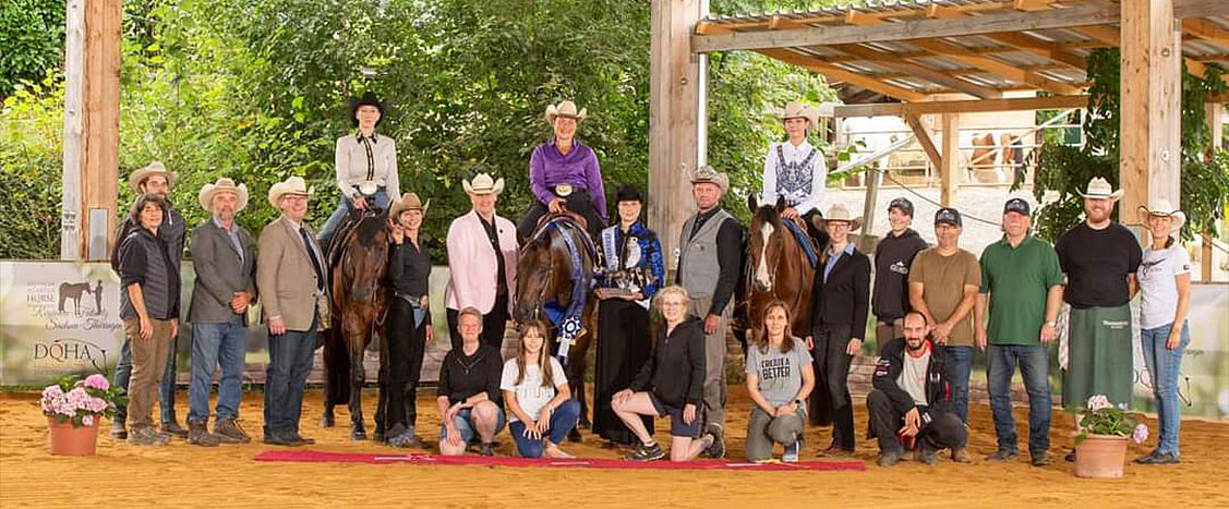 Großes Gruppenfoto bei einer Westernreitveranstaltung: Reiterinnen und Reiter mit Cowboyhüten, mehrere gesattelte Pferde, Funktionäre und Helfer posieren gemeinsam unter einem Holzunterstand; einige Personen halten Pokale, im Hintergrund sind Bäume und Stallgebäude zu sehen.