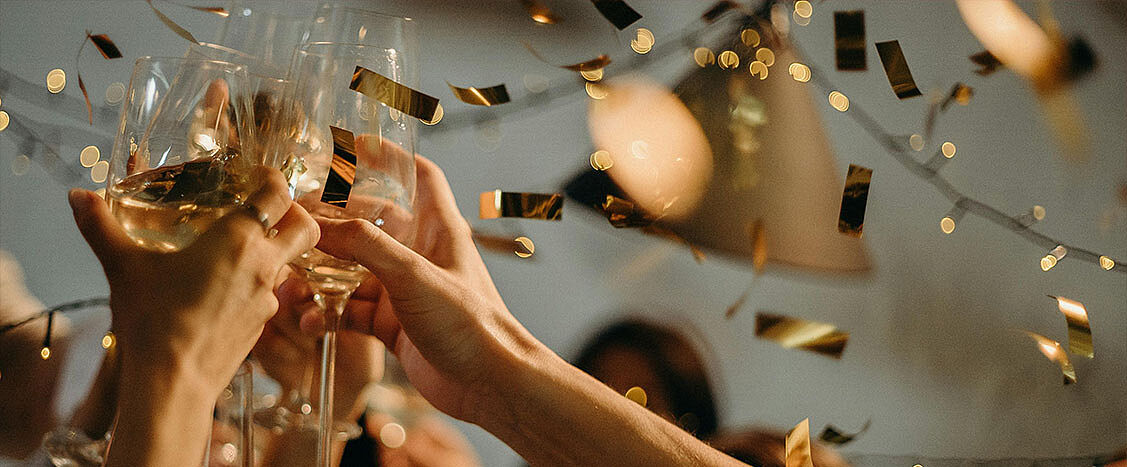 Mehrere Hände stoßen mit gefüllten Weingläsern an, während goldenes Konfetti durch die Luft fällt. Warmes Licht und unscharfe Lichter im Hintergrund vermitteln eine ausgelassene, festliche Stimmung.