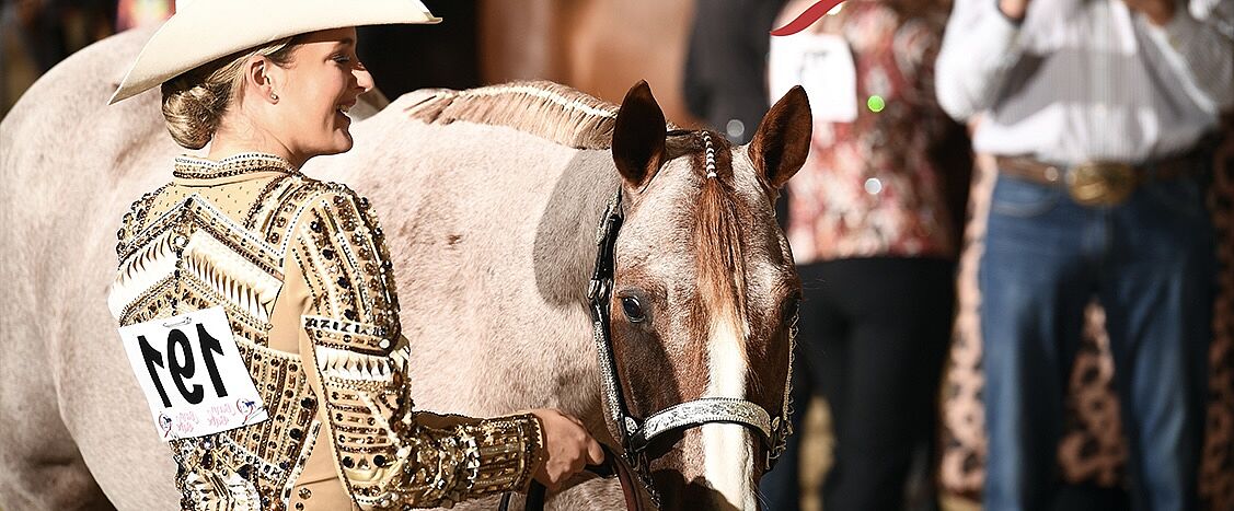 Eine Reiterin mit Cowboyhut und verzierter Turnierkleidung hält ein gesatteltes Pferd am Zügel; auf ihrem Rücken ist die Startnummer „191“ befestigt, im Hintergrund stehen mehrere Personen.