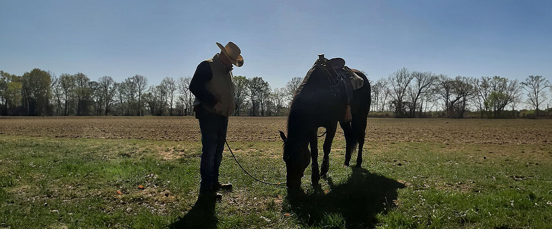 Eine Person mit Cowboyhut steht auf einer Wiese neben einem gesattelten dunklen Pferd, das mit gesenktem Kopf grast; im Hintergrund ein Feld und Bäume.