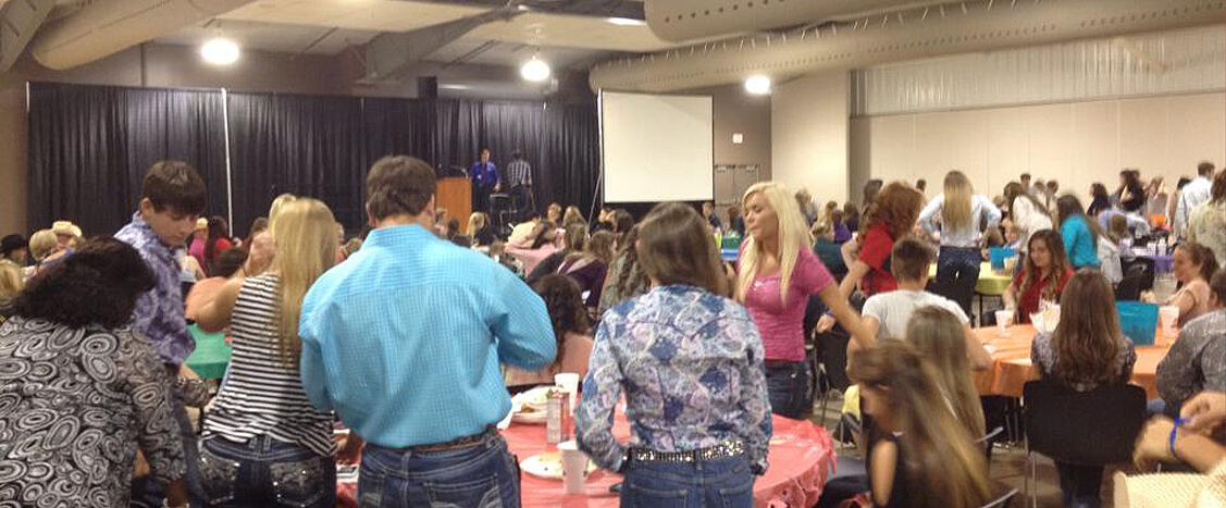 Großer Saal mit vielen stehenden und sitzenden Personen an runden Tischen, teilweise in Westernkleidung; im Hintergrund eine Bühne mit Rednerpult und Leinwand. Bunte Tischdecken und dichtes Gedränge vermitteln eine Veranstaltung mit großem Publikumsandrang.
