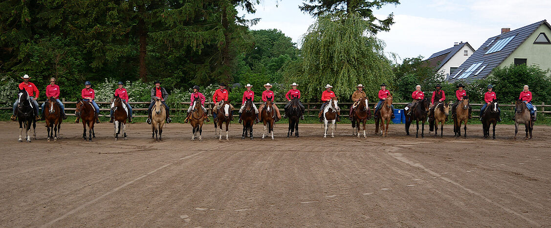 Eine große Gruppe von Reiterinnen und Reitern in roten Shirts sitzt auf ihren Pferden in einer geraden Linie auf einem Reitplatz. Die Szene wirkt geordnet und gemeinschaftlich, eingerahmt von Bäumen und Wohnhäusern im Hintergrund.
