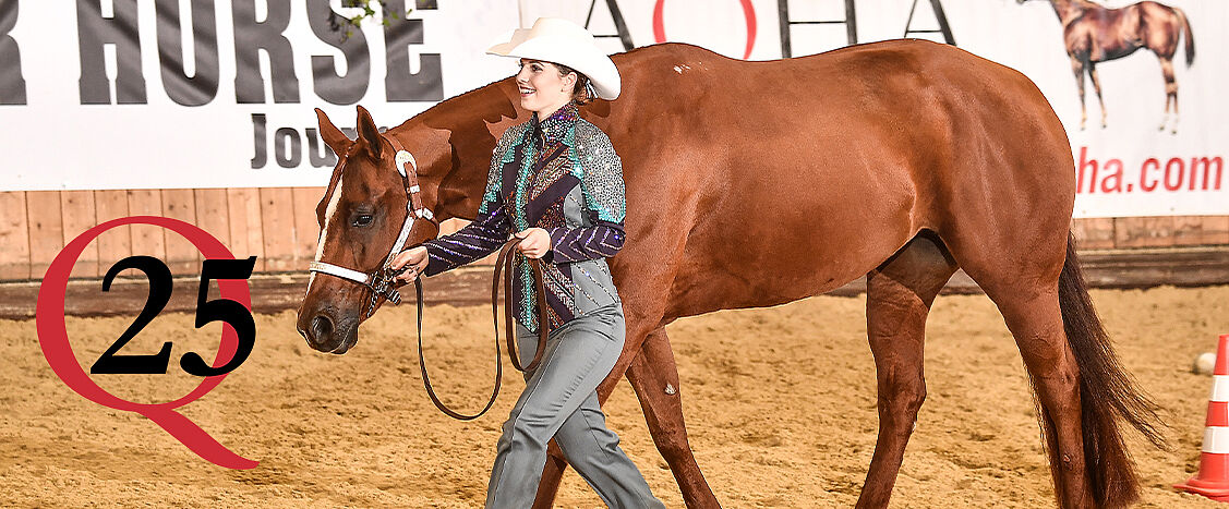 Reiterin in Westernkleidung führt ein braunes Quarter Horse im Schritt durch eine Reithalle; sie trägt einen weißen Cowboyhut, im Hintergrund sind Hallenbande, Schriftzüge und ein rotes „Q25“-Logo zu sehen.