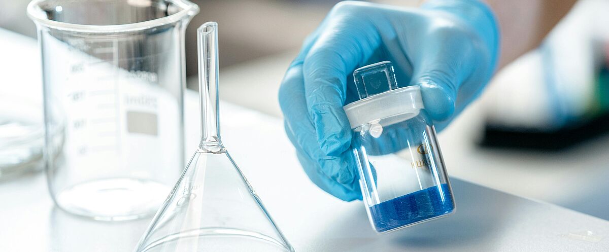 Eine Hand mit blauem Einweghandschuh hält ein kleines Glasgefäß mit blauer Flüssigkeit; daneben stehen ein Becherglas und ein Glastrichter auf einer Arbeitsfläche.