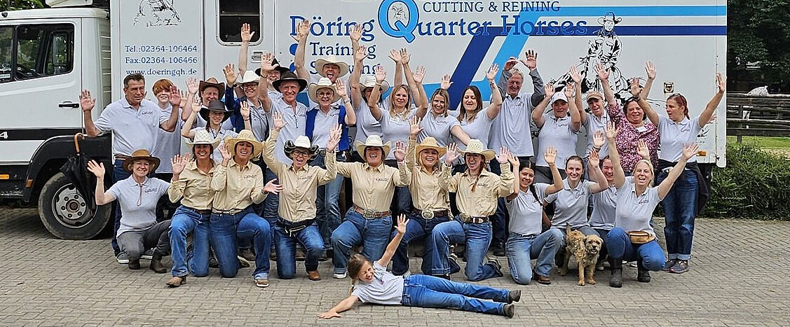 Große Gruppe von Personen steht und kniet vor einem Transporter und hebt die Hände; einige tragen Cowboyhüte, eine Person liegt vorne auf dem Boden, daneben ein kleiner Hund. Auf dem Transporter steht der Text „Döring Quarter Horses – Cutting & Reining – Training & Sales“ sowie Telefonnummern und eine Website.