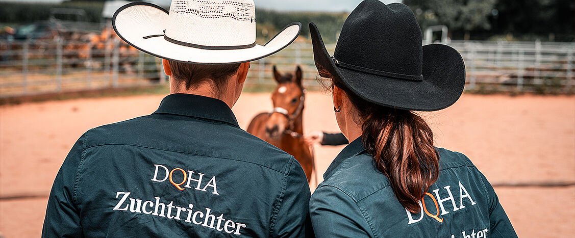 Zwei Personen stehen mit dem Rücken zur Kamera in einer Reitbahn. Sie tragen Westernhüte und Hemden mit der Aufschrift „DQHA Zuchtrichter“. Vor ihnen steht ein junges Pferd, das von einer weiteren Person geführt wird. Die Szene spielt im Freien auf sandigem Boden, im Hintergrund sind Zäune und eine Reitanlage zu sehen.