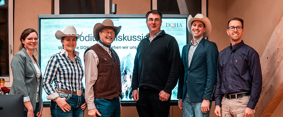 Sechs Personen stehen nebeneinander vor einem Bildschirm mit der Aufschrift „Podiumsdiskussion“ und dem Logo der Deutschen Quarter Horse Association. Einige tragen Westernhüte; der Raum wirkt wie ein Veranstaltungs- oder Tagungsraum.