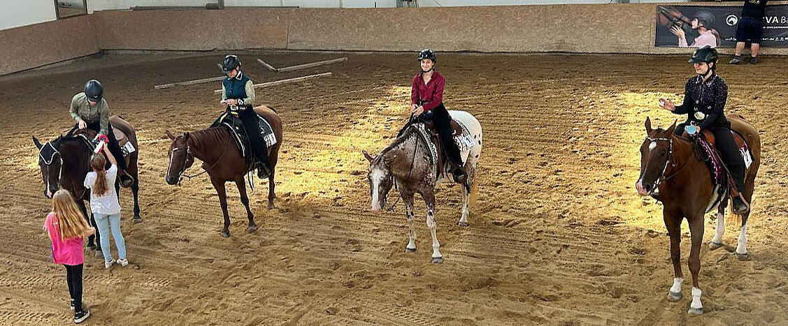 Mehrere junge Reiterinnen sitzen mit Schutzhelmen auf Westernpferden in einer Reithalle mit Sandboden; zwei Kinder stehen in der Mitte und sprechen mit einer Reiterin, während die übrigen Pferde ruhig im Kreis warten.