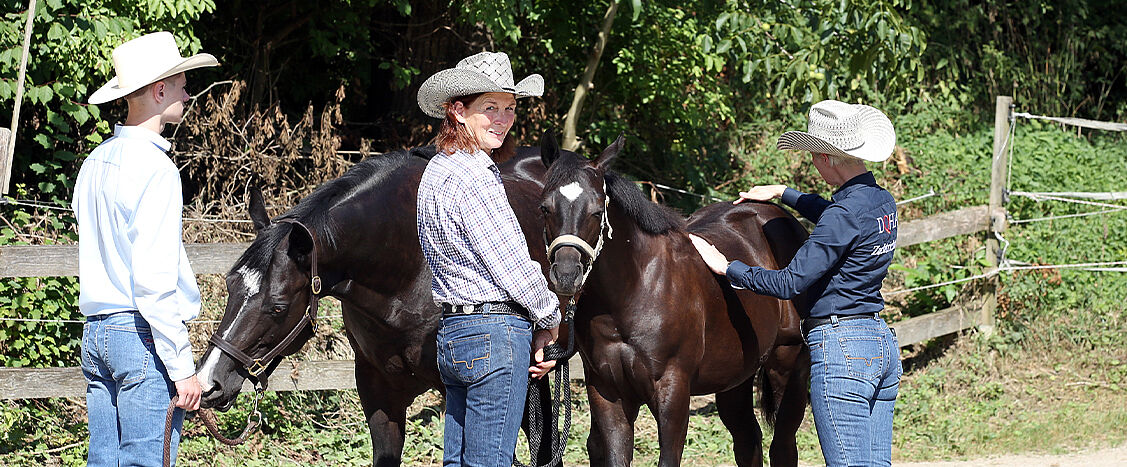 Drei Personen mit Cowboyhüten stehen mit zwei schwarzen Pferden an einem Holzzaun im Grünen.