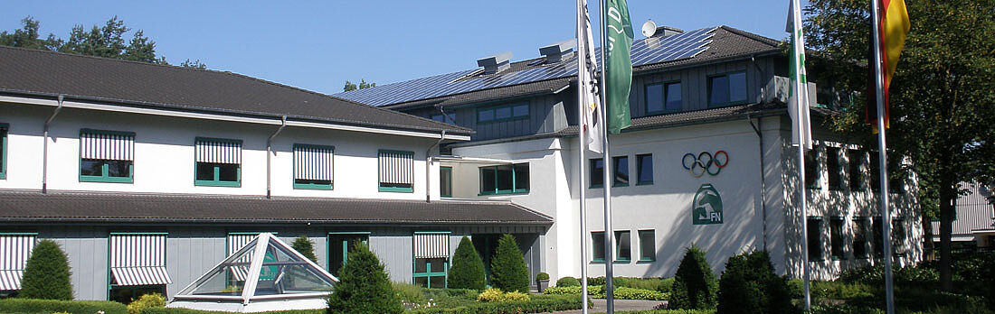 Ein modernes, helles Gebäude mit gepflegter Grünanlage und mehreren Fahnenmasten liegt ruhig im Sonnenlicht. Klare Architektur, saubere Linien und die eingebettete Lage im Grünen vermitteln einen ordentlichen, institutionellen Eindruck.