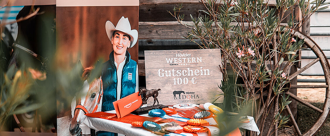 Dekorierter Tisch mit Preisschleifen und Werbematerial; daneben ein Schild mit dem Text „Höveler WESTERN Gutschein 100 €“ und „DQHA Zuchtschauen“. Im Hintergrund ein Plakat mit einer Person im Westernhut neben einem Pferd sowie Holzwand und Pflanzen.