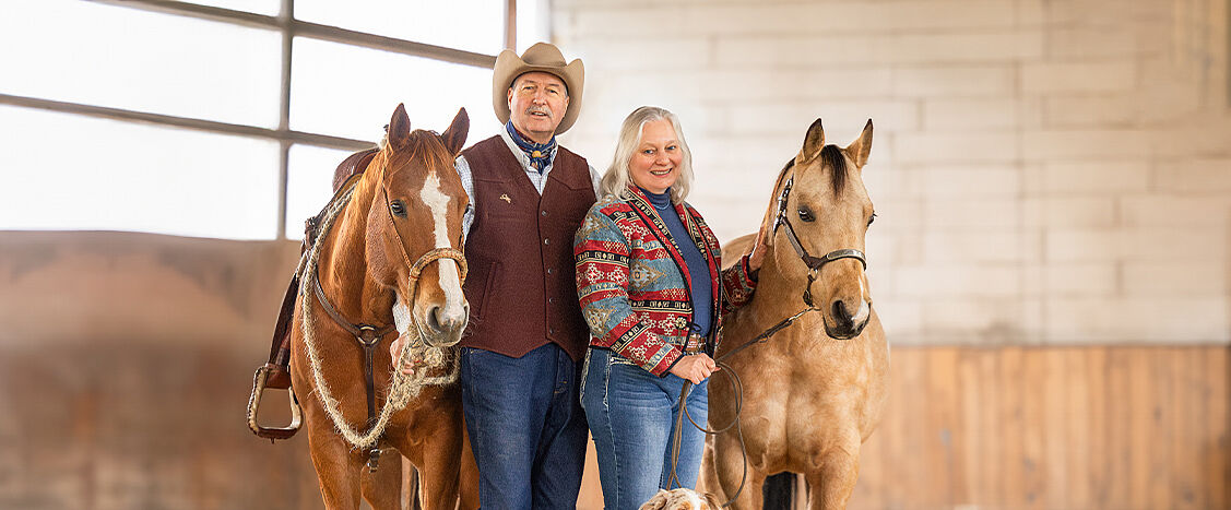 Zwei Personen stehen in einer Reithalle zwischen zwei Pferden; vor ihnen sitzt ein Hund im Sand.