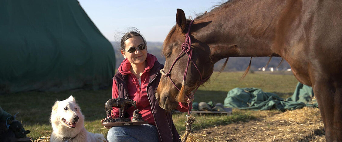 Eine Person sitzt im Freien neben einem braunen Pferd und hält eine kleine Trophäe; daneben liegt ein weißer Hund auf Stroh.