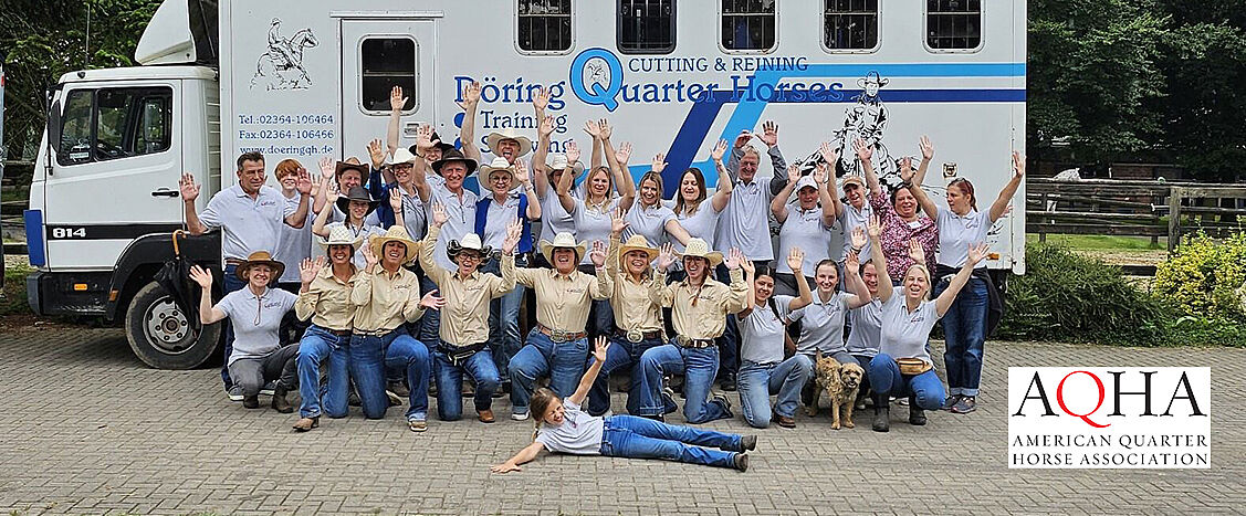 Große Gruppe von Menschen in Westernkleidung steht vor einem Trainings-LKW, viele mit erhobenen Armen und fröhlichen Gesichtern; einige knien oder sitzen vorne, ein Hund liegt in der Mitte auf dem Boden.