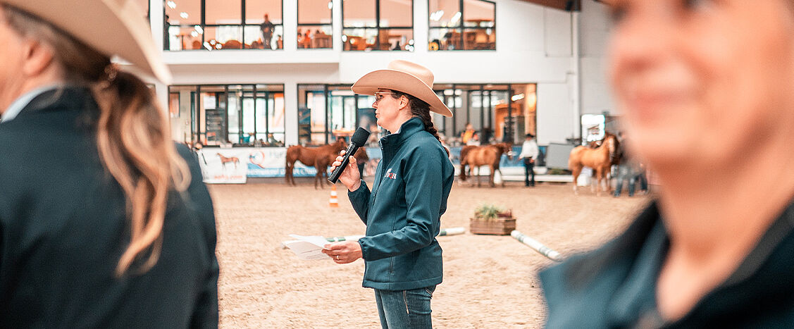 Eine Sprecherin mit Cowboyhut hält ein Mikrofon und spricht in einer Reithalle, während im Hintergrund Pferde und Zuschauer zu sehen sind. Die Szene zeigt eine Moderation oder Siegerehrung im Rahmen einer Pferdesportveranstaltung.