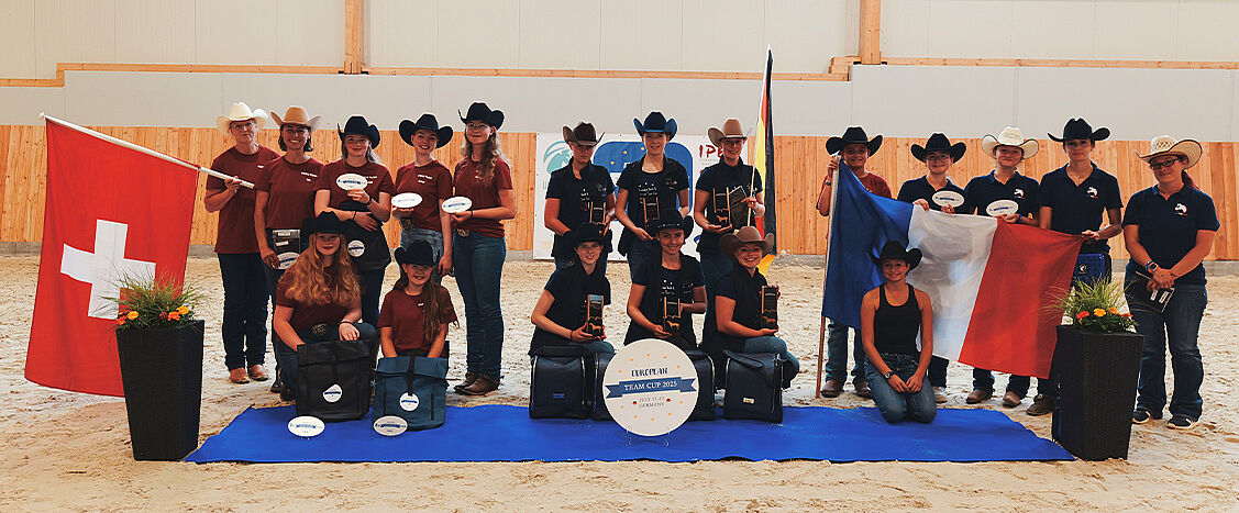 Gruppenfoto von Personen in Reithalle mit Schweizer und französischer Flagge, einige tragen Cowboyhüte, vor ihnen stehen Trophäen und Taschen auf blauem Bodenbelag