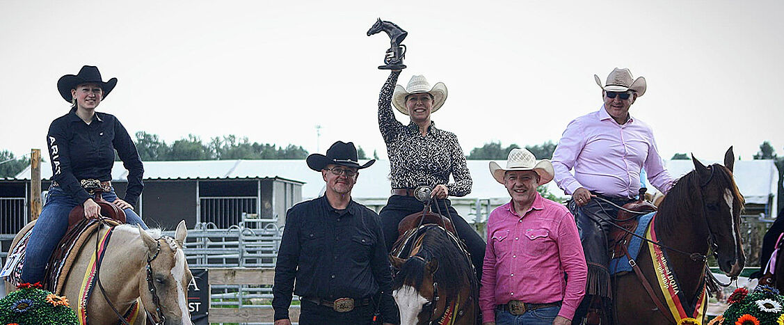 Siegerehrung im Westernreitsport auf einem Reitplatz im Freien. Mehrere Reiterinnen und Reiter in Cowboyhüten sitzen auf gesattelten Pferden oder stehen davor. In der Mitte hebt eine Reiterin auf dem Pferd eine Trophäe über den Kopf. Die Pferde tragen Siegerschleifen in den Farben Schwarz-Rot-Gold. Im Hintergrund sind Stallgebäude, Absperrungen und Zuschauerbereiche zu sehen.