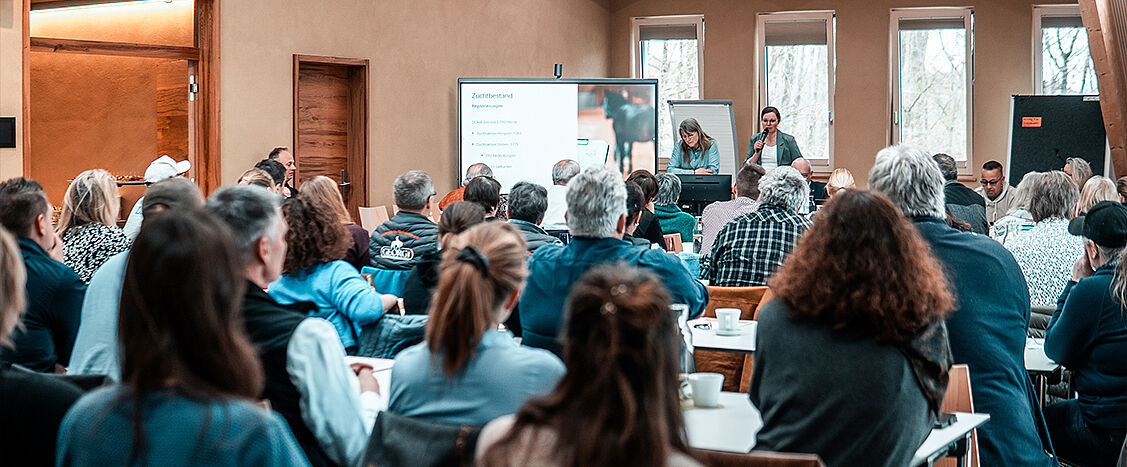 Großer Versammlungsraum mit vielen sitzenden Personen an Tischen, vorne zwei Personen an einem Rednerpult. Hinter ihnen eine Leinwand mit Präsentation, Fenster und Holzdecke sind sichtbar.