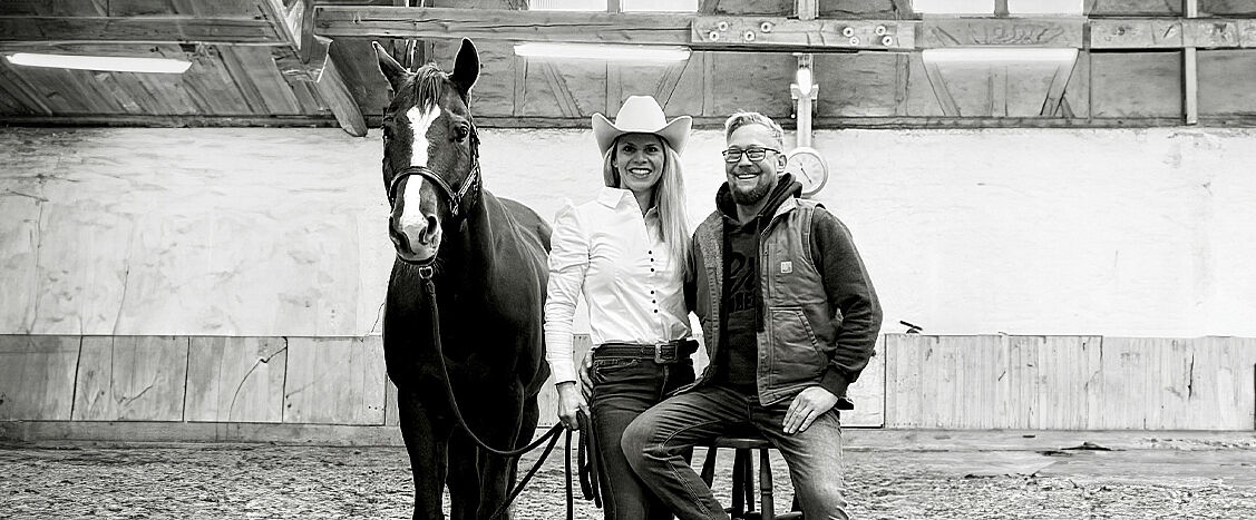 Eine Frau im Westernhut und ein Mann sitzen in einer Reithalle neben einem gesattelten Pferd und lächeln in die Kamera. Die Szene ist in Schwarz-Weiß gehalten und wirkt ruhig und vertraut.