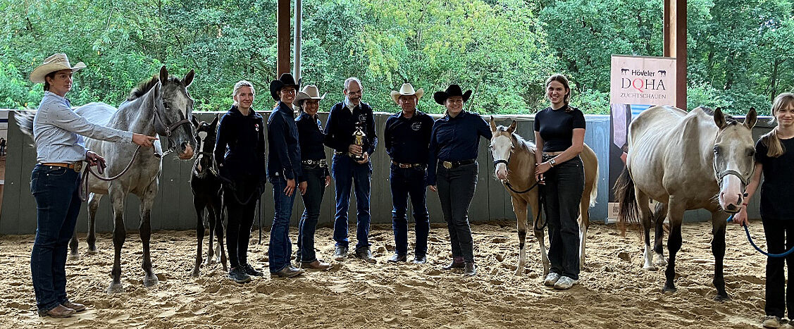Eine Gruppe von Züchtern, Richtern und Helfern steht mit mehreren Pferden in einer überdachten Reithalle. Im Hintergrund sind Banner der DQHA-Zuchtschau zu sehen.