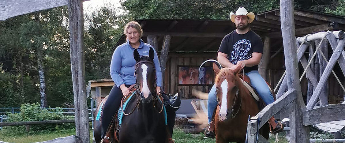 Zwei Reiter sitzen entspannt auf ihren Pferden vor einer rustikalen Holzkulisse, eingerahmt von Bäumen und einem einfachen Unterstand. Die Szene wirkt ruhig und vertraut, mit einem natürlichen, ländlichen Western-Ambiente.