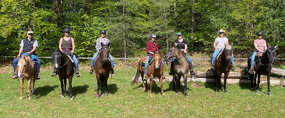 Sieben Personen sitzen nebeneinander auf Pferden auf einer Wiese vor einem Waldrand; die Pferde stehen still und die Reitenden tragen Helme oder Hüte.