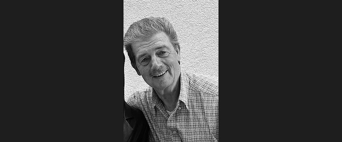 Schwarzweiß-Porträt eines lächelnden Mannes vor heller Wand.