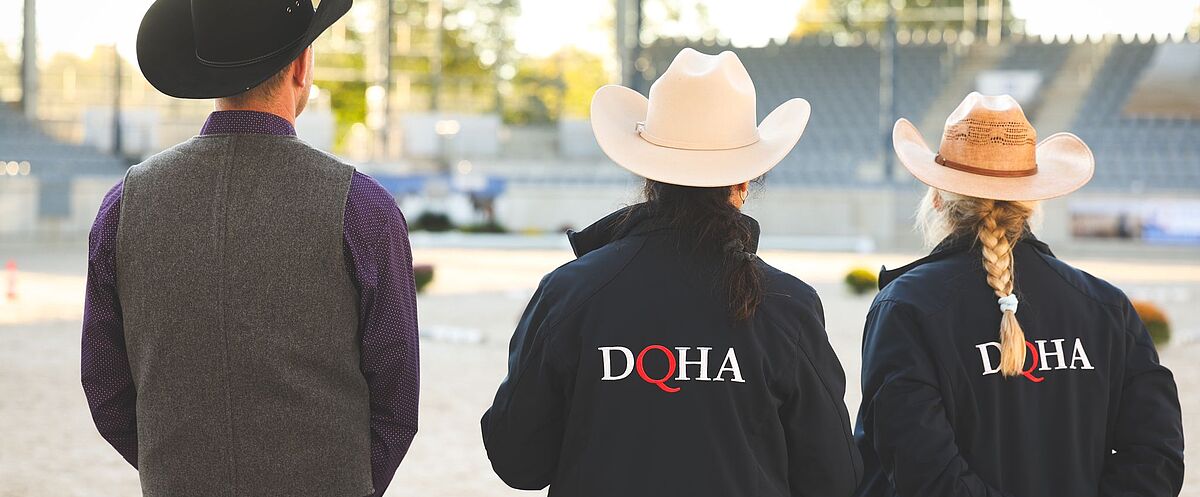 Drei Personen mit Cowboyhüten stehen von hinten in einer Reithalle und blicken auf den Showring. Zwei tragen Jacken mit dem „DQHA“-Logo und beobachten gemeinsam das Geschehen.