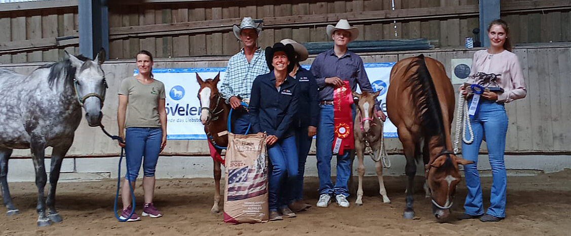 Mehrere Personen stehen in einer Reithalle zwischen Stuten und Fohlen bei einer Siegerehrung; einige Pferde tragen rote Siegerschleifen, im Hintergrund hängt ein Banner mit der Aufschrift „Höveler“.