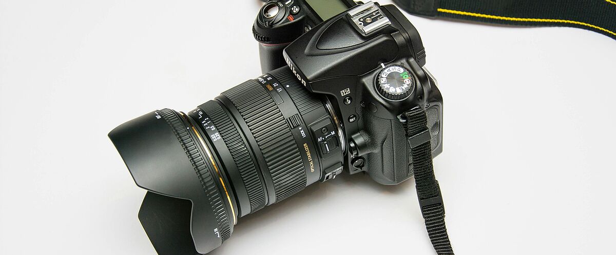 Schwarze digitale Spiegelreflexkamera mit angesetztem Zoomobjektiv und Gegenlichtblende, daran befestigt ein Kameragurt, liegend auf weißem Hintergrund.