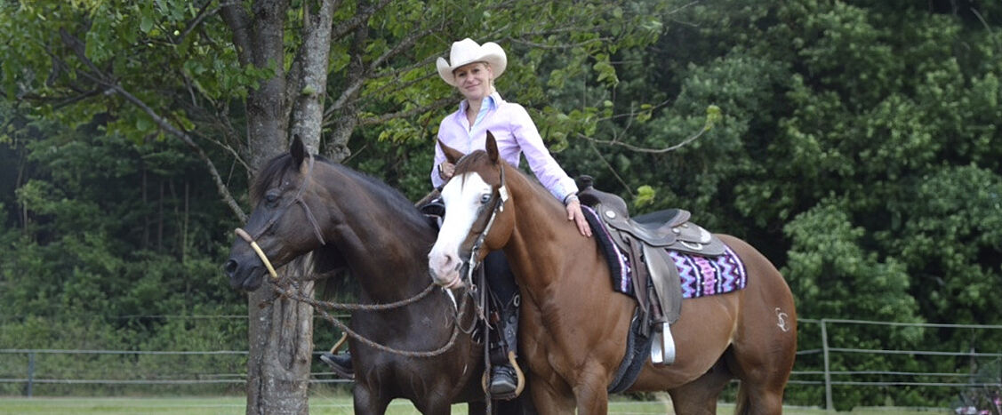 Reiterin mit Cowboyhut sitzt auf einem braunen Pferd, daneben ein weiteres dunkles Pferd, im Grünen.