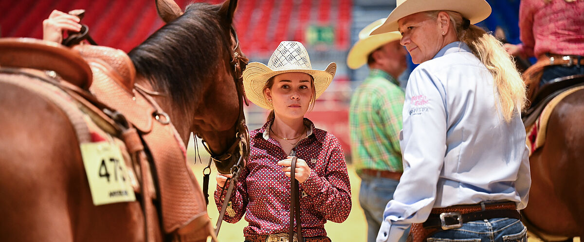 Mehrere Personen mit Cowboyhüten stehen mit gesattelten Pferden in einer Reithalle; im Vordergrund hält eine Person die Zügel eines Pferdes, eine weitere Person steht daneben. Oben links ist das Logo „AMERICANA 2023“ zu sehen, im Hintergrund rote Tribünensitze.