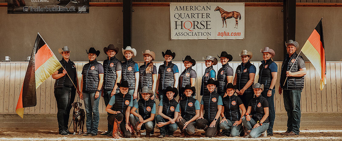 Gruppe von Personen in Reitbekleidung mit zwei deutschen Flaggen in einer Reithalle vor einem Schild der American Quarter Horse Association