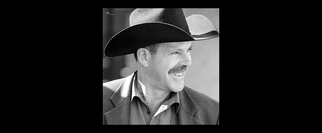 Schwarz-weißes Porträt eines Mannes mit Cowboyhut und Schnurrbart, der lächelnd zur Seite blickt. Er trägt ein Sakko über einem Hemd, der Hintergrund ist dunkel und ruhig gehalten.