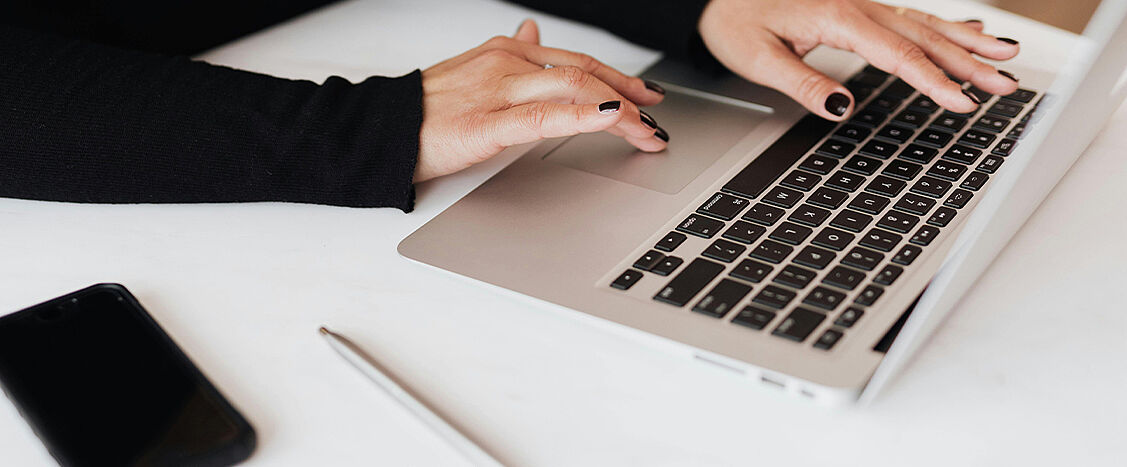 Zwei Hände tippen auf der Tastatur eines Laptops auf einem weißen Schreibtisch. Daneben liegen ein Smartphone und ein Stift.