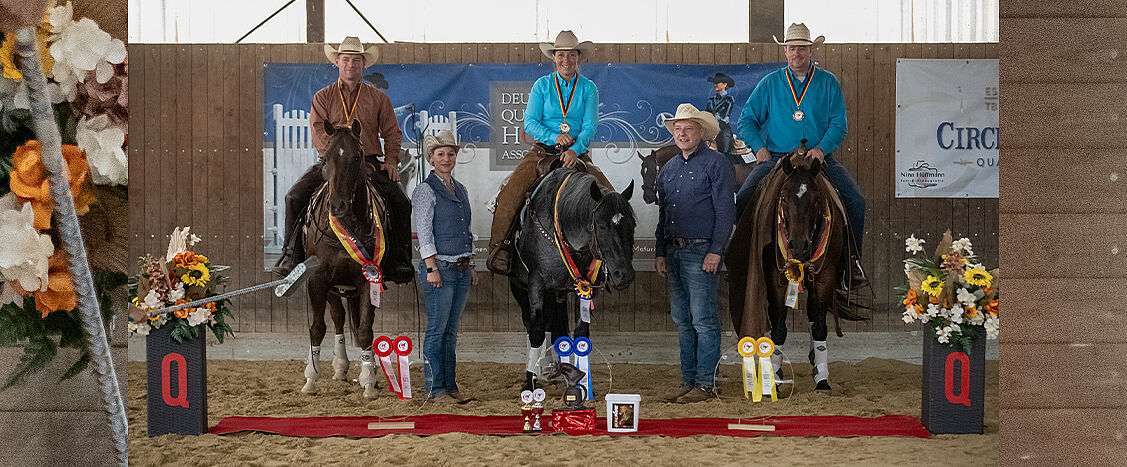 Siegerehrung in einer Reithalle: Drei Reiter sitzen auf ihren Pferden und tragen Medaillen um den Hals, flankiert von zwei weiteren Personen am Boden. Vor den Pferden liegen farbige Schleifen und Pokale auf einem roten Teppich. Links und rechts stehen dekorative Blumengestecke mit schwarzen Sockeln und rotem „Q“-Logo. Im Hintergrund sind Banner der Deutschen Quarter Horse Association und weiterer Sponsoren an der Hallenwand angebracht.